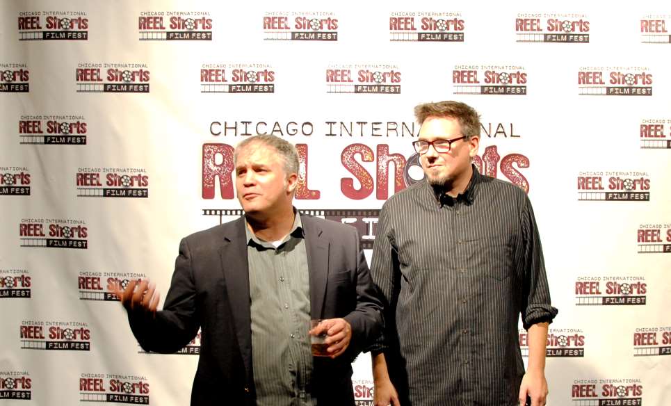 Two men at an event: one gesturing, the other smiling. Background: 