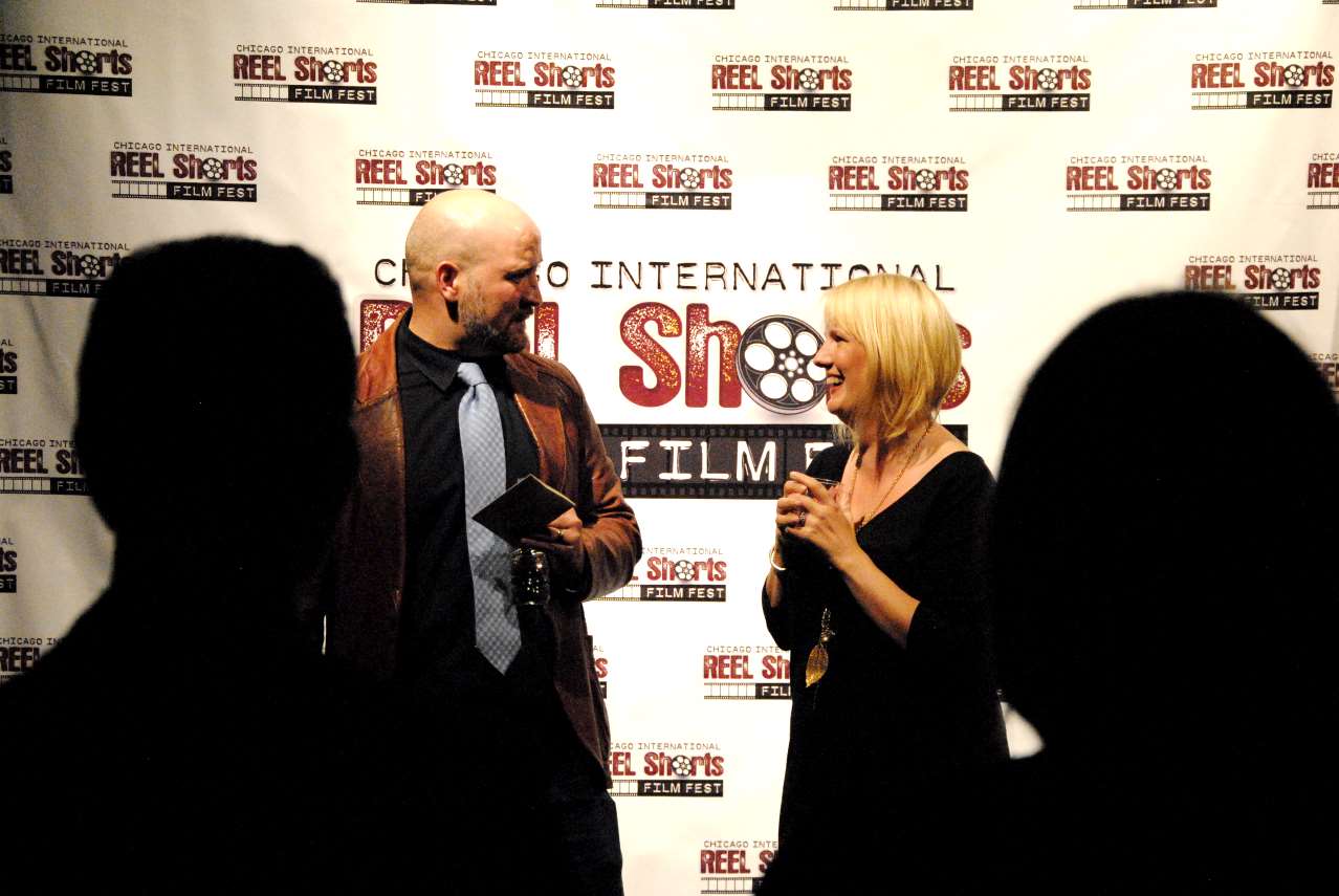 Man accepting award, woman holding microphone at film festival. Red backdrop.