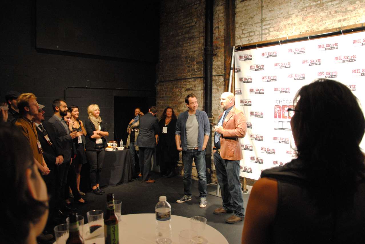 People at an event, listening to two men speak in front of a photo backdrop.