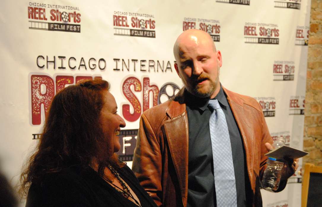 Man in brown jacket and woman pose at Chicago film festival, red backdrop.
