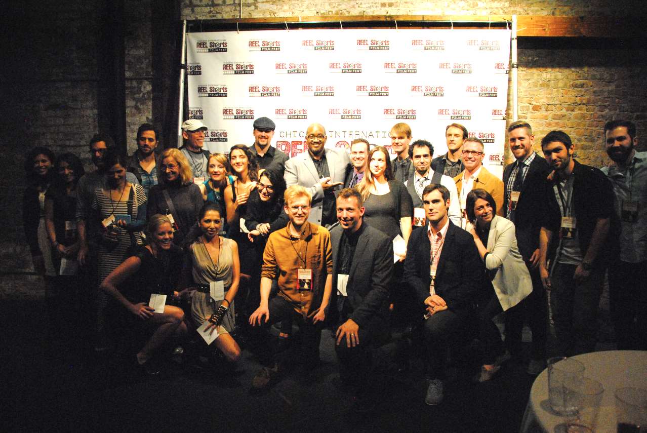Group of people posing in front of a backdrop, likely at an event. Dark setting, mix of casual and formal attire.