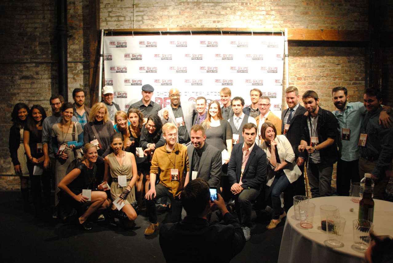 Group posing for photo; people in a room with a branded backdrop. Some are kneeling. Mixed ages, diverse clothing.