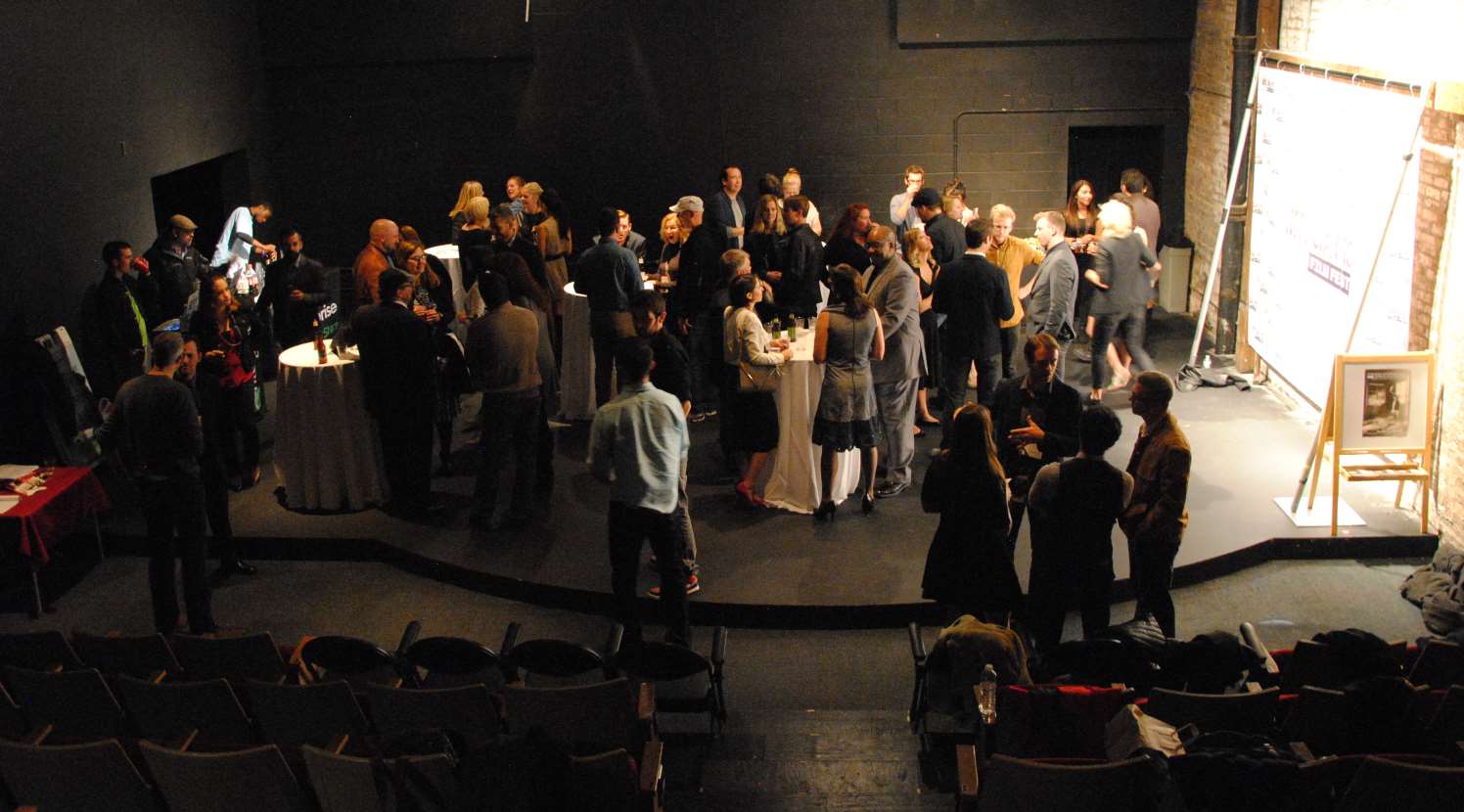 People mingling at an event in a theater. Dark walls, cocktail tables, and a projected image on a screen.