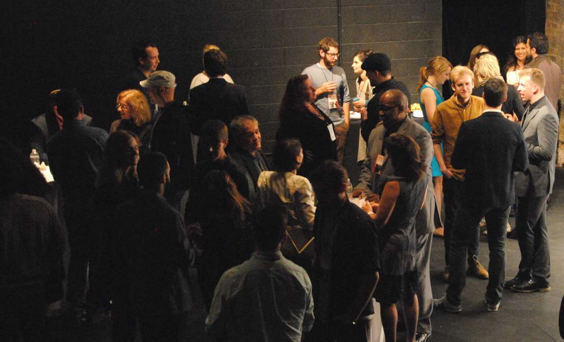 A crowd of people socializing in a dimly lit room. Some are talking, and others are looking around.