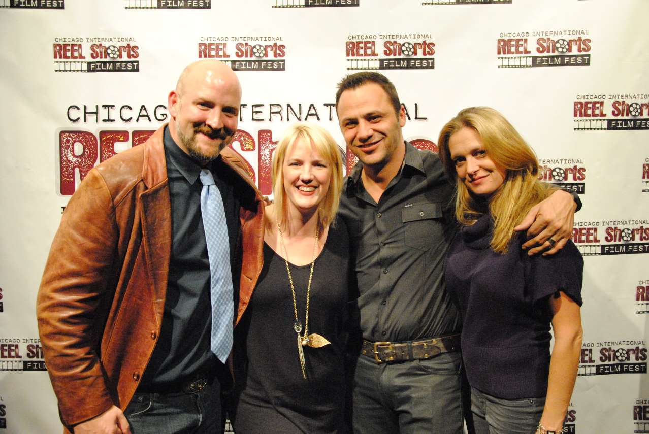 Four people smiling at a film festival. Woman in blue embraces man in a black shirt; man in leather jacket stands left.