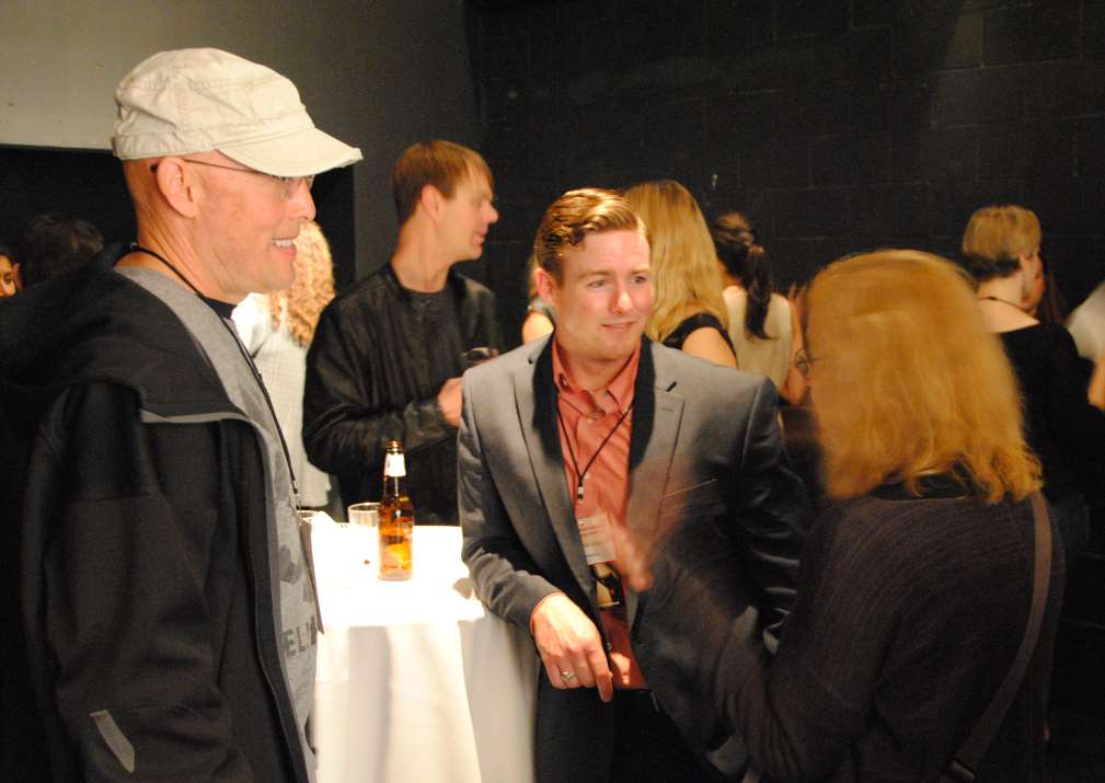 People at an event, conversing. Man in a blazer talks with a woman. Others stand around. Low lighting.