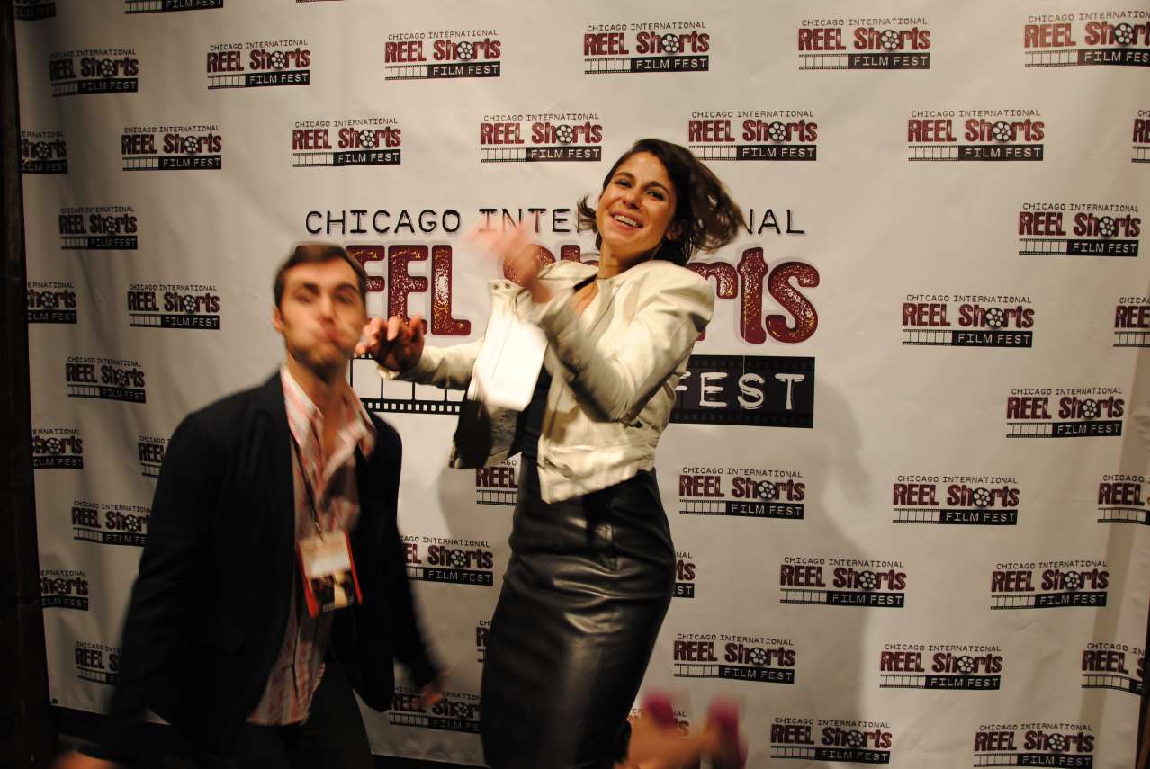 A woman jumps excitedly near a man, both at Chicago Reel Shorts Fest. Background features the festival logo.