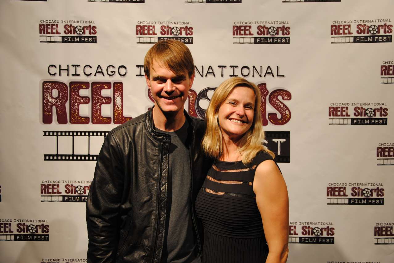 A man and woman smiling at a film festival backdrop. Man in a black jacket, woman in a black dress.