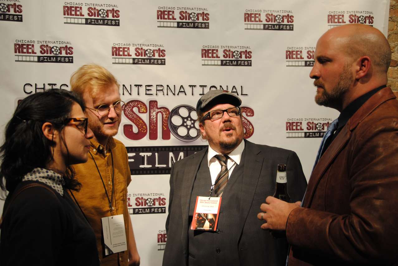 Four people pose for a photo at a film festival. White backdrop with event logo. Man with beard holding a beer.