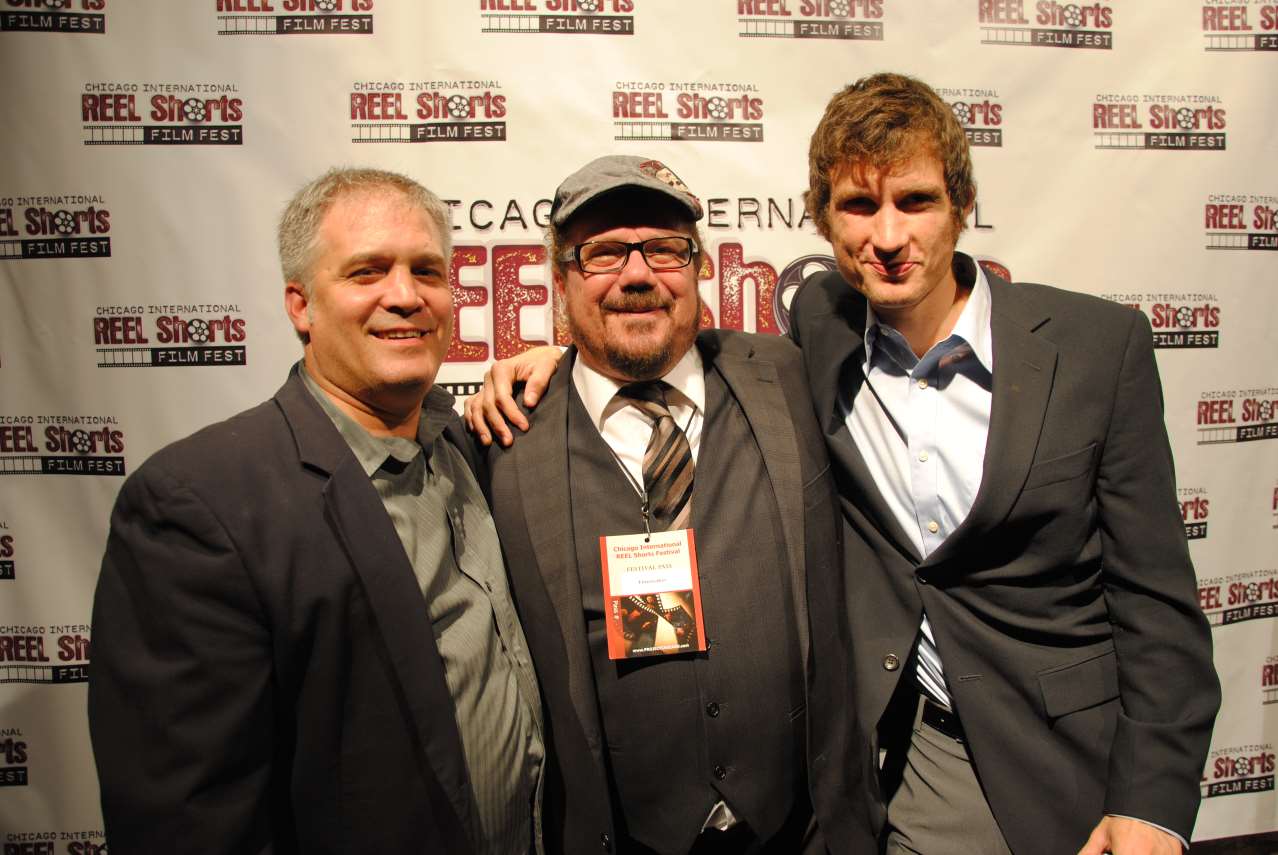 Three men at a film festival posing for a photo. One is wearing a hat, they're all smiling.