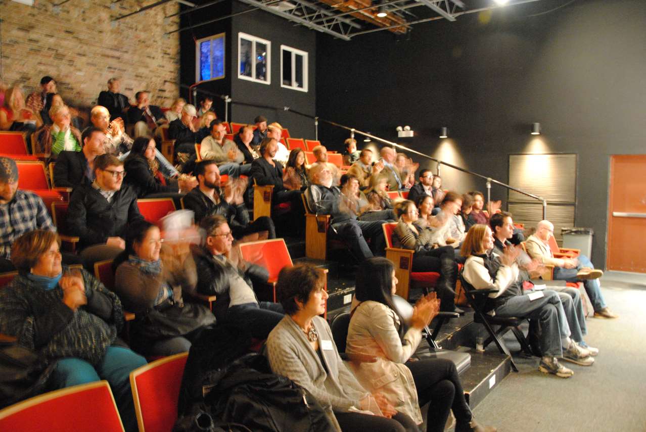 Audience applauds in a small theater with red seats, a stage-left doorway, and a black wall.