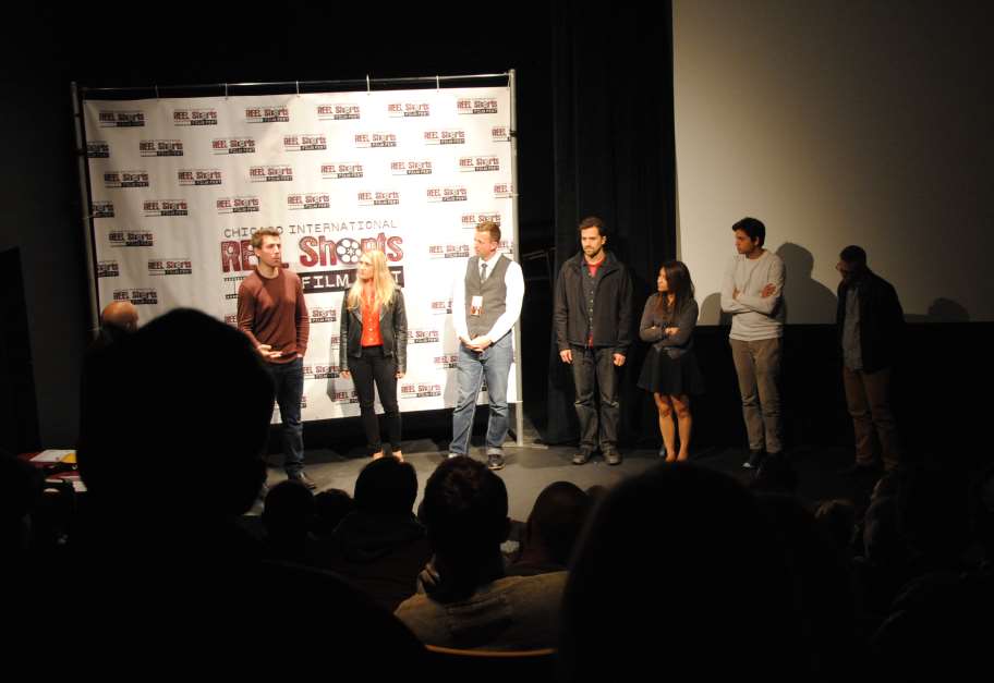 Panel of people on stage at film event.  Backdrop with logos. Audience silhouettes in foreground.