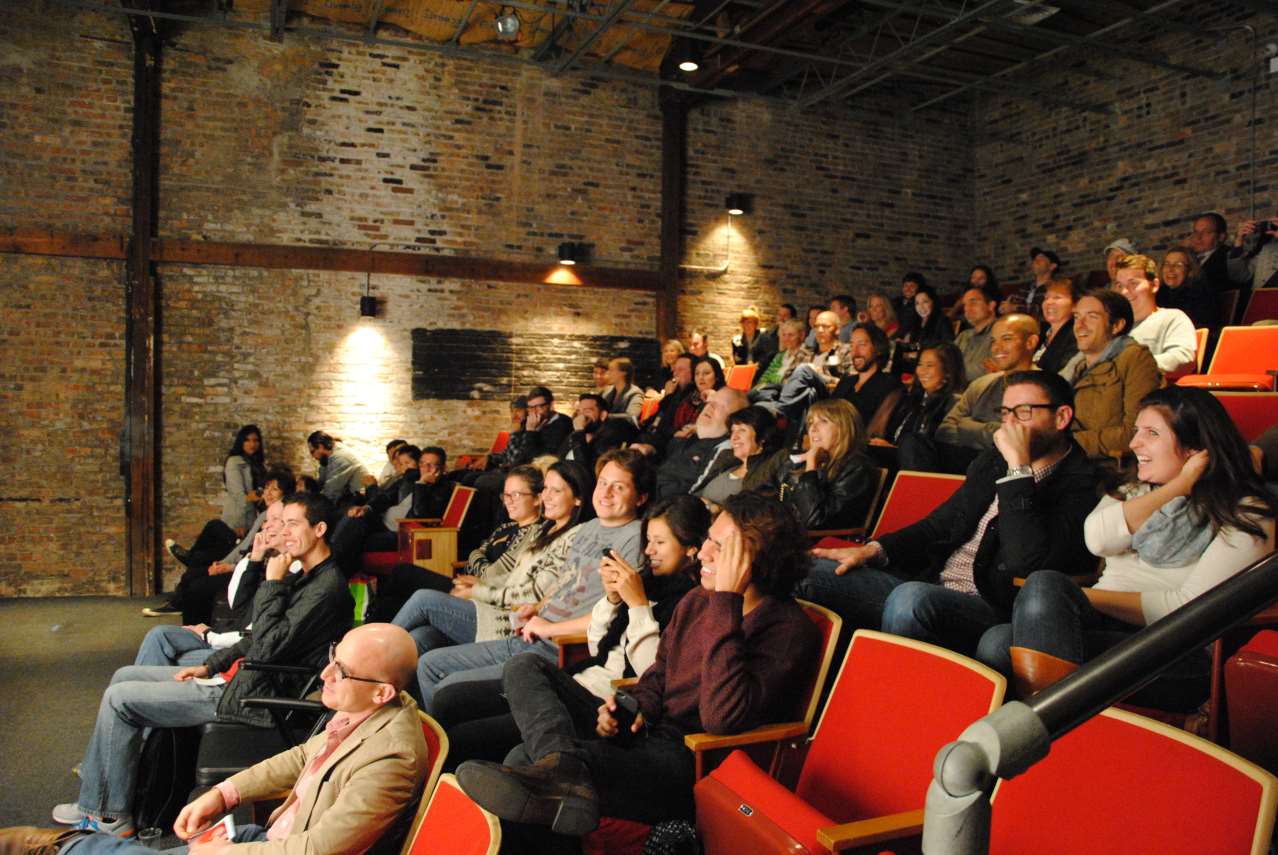 Audience in red theater seats, watching a performance in a brick-walled venue.