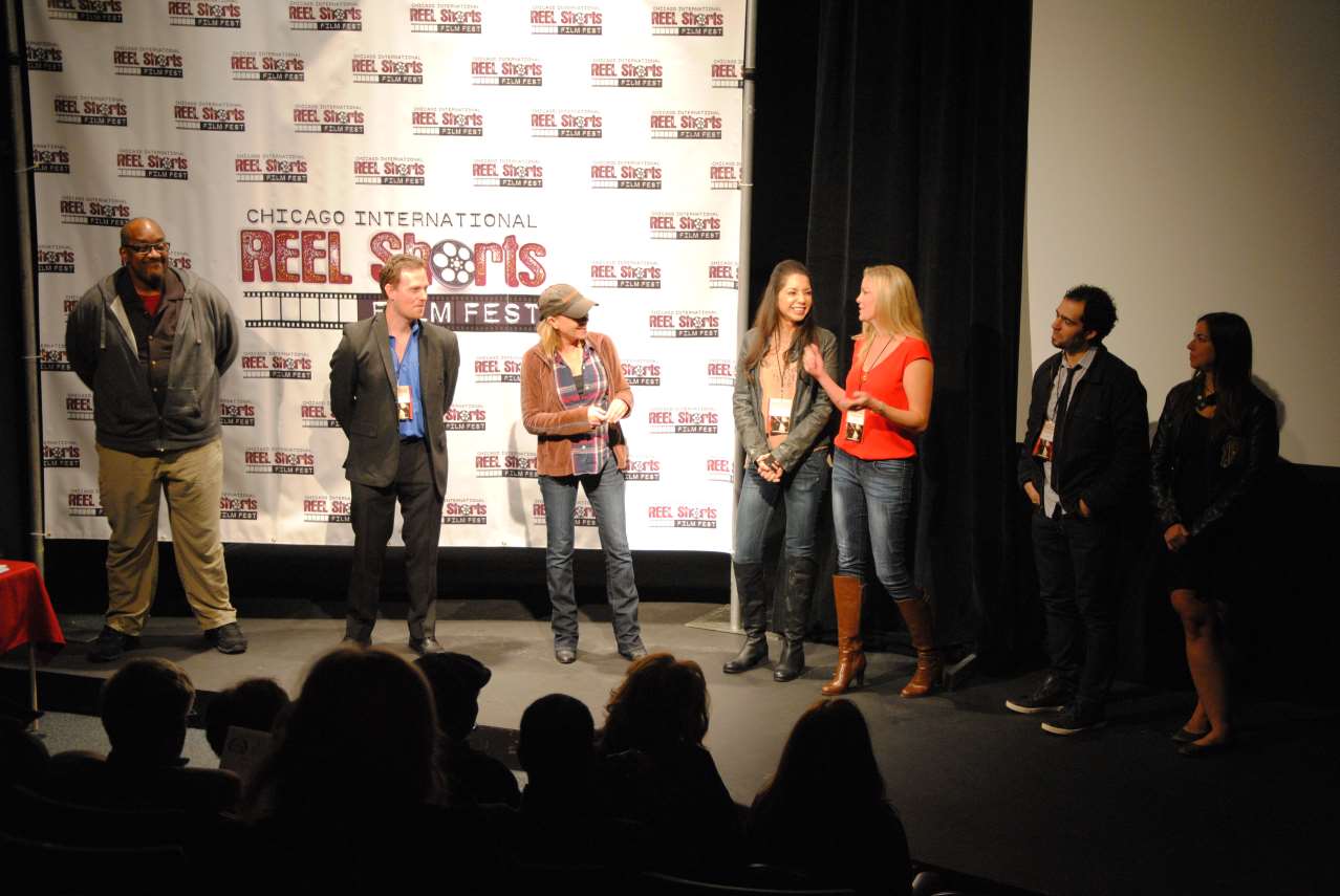 People on stage at a film festival; two women with microphones, others standing, with audience in front.
