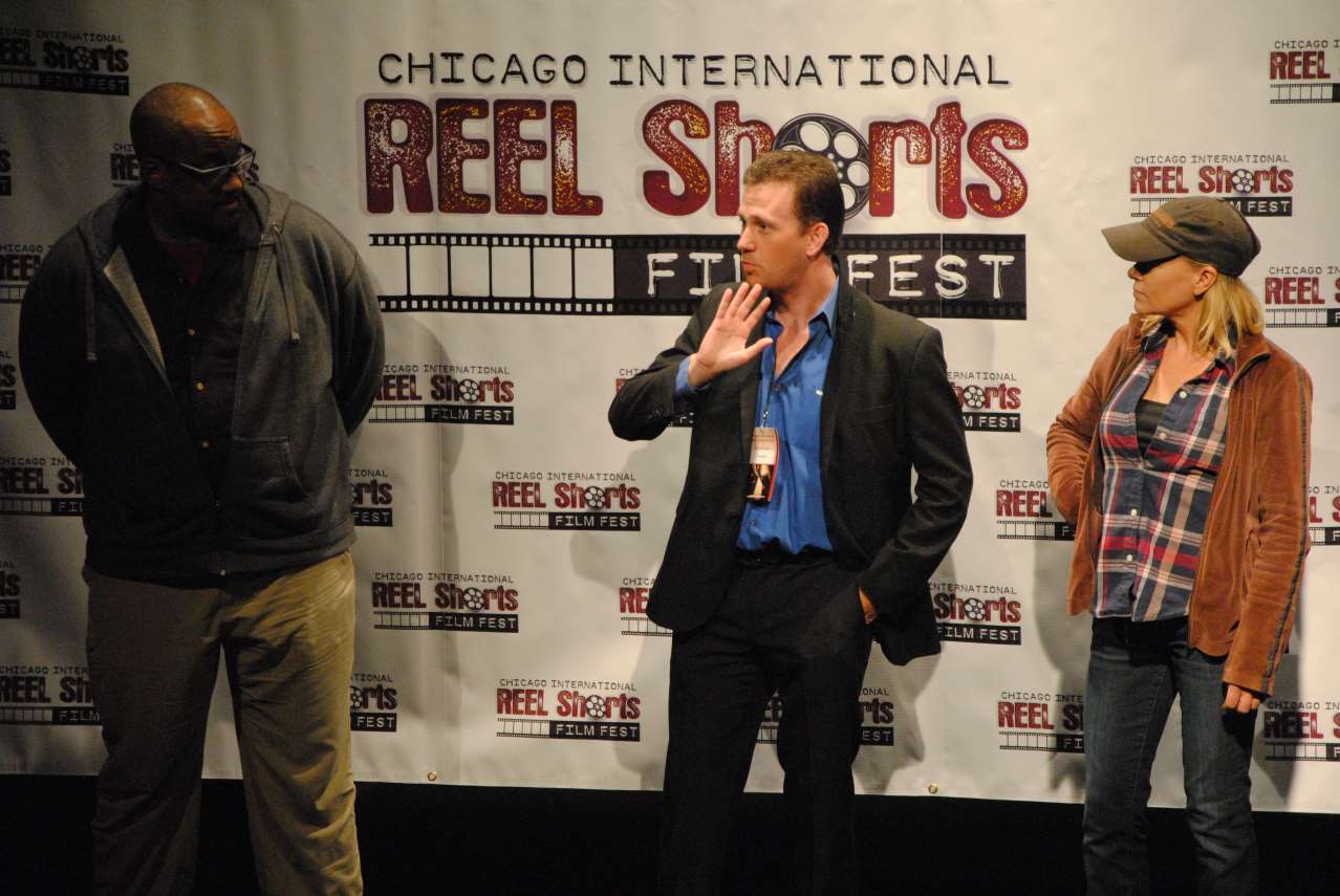 Three people onstage at the Chicago Reel Shorts Film Fest. A man gestures, while two others stand beside him.