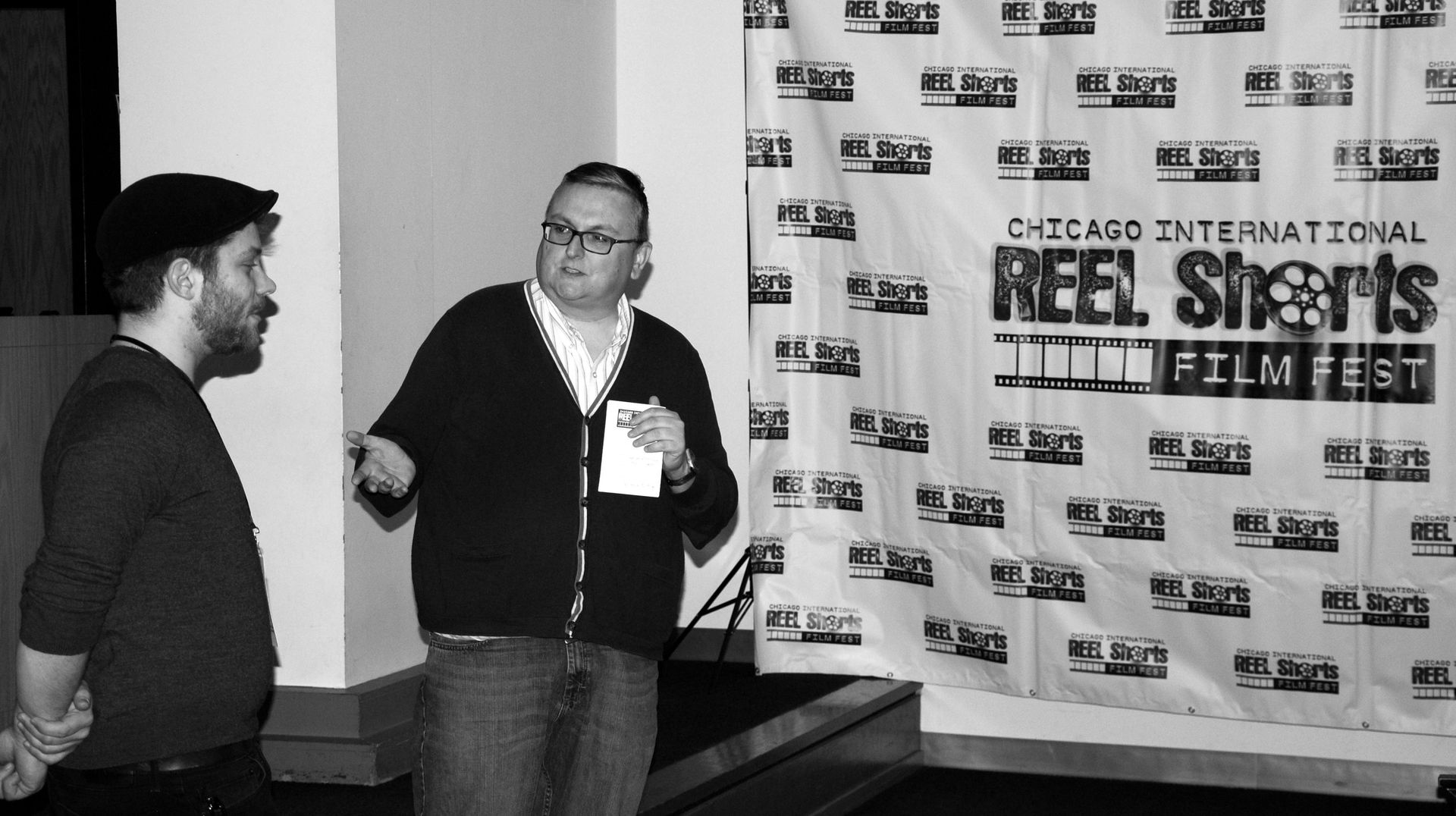 Two men in front of a banner for the Oakland International Reel Shorts Film Fest; one speaks, gesturing.