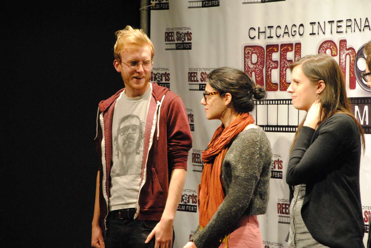 Three people at a film festival. A man in a red hoodie talks to a woman in glasses and a scarf, another woman listens.