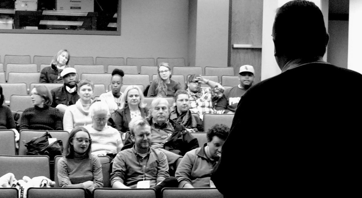 Audience seated in rows listens to a speaker in a lecture hall. Black and white photo.