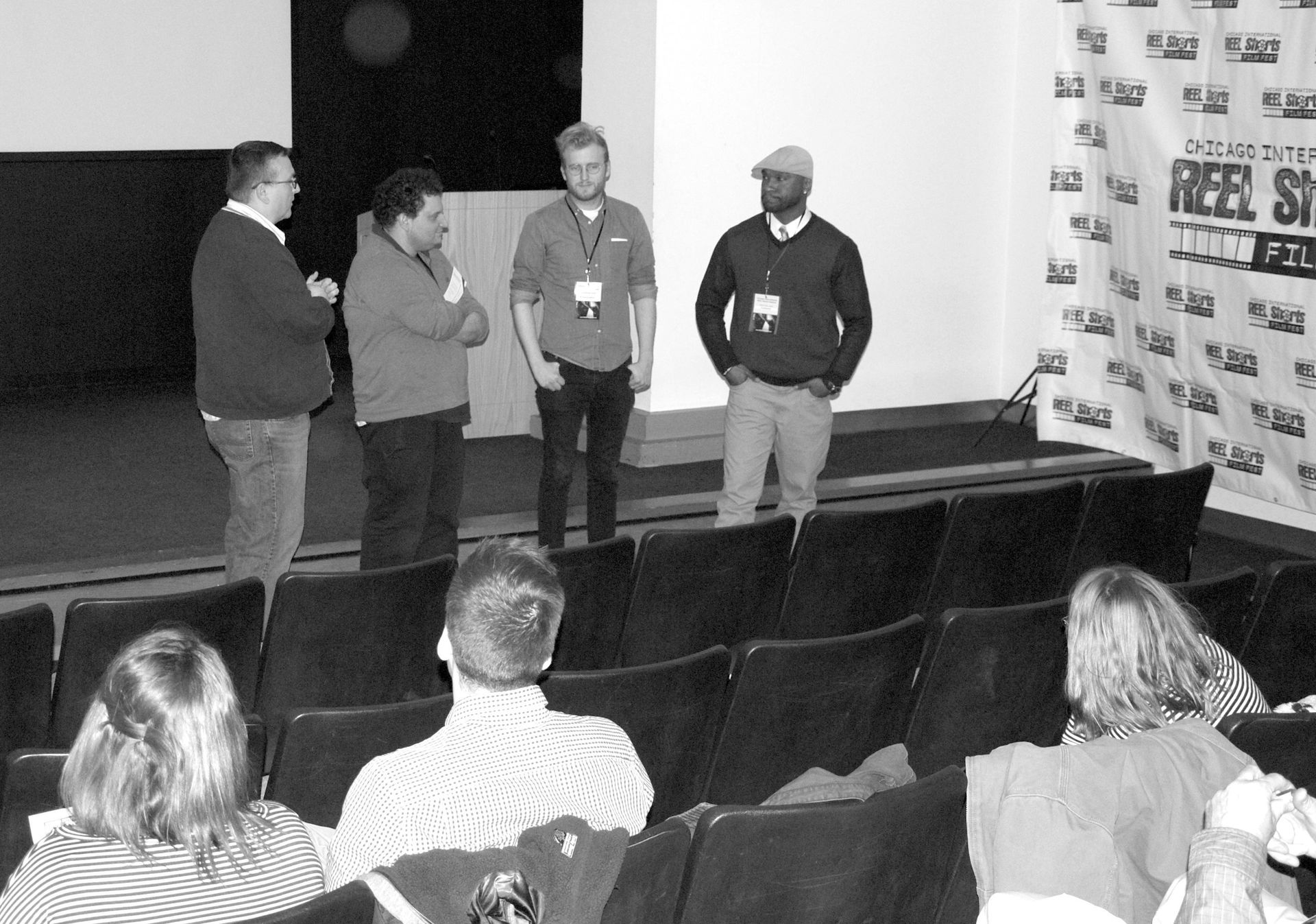 Panel of four men on a stage, speaking to an audience seated in rows of chairs; banners in the background.