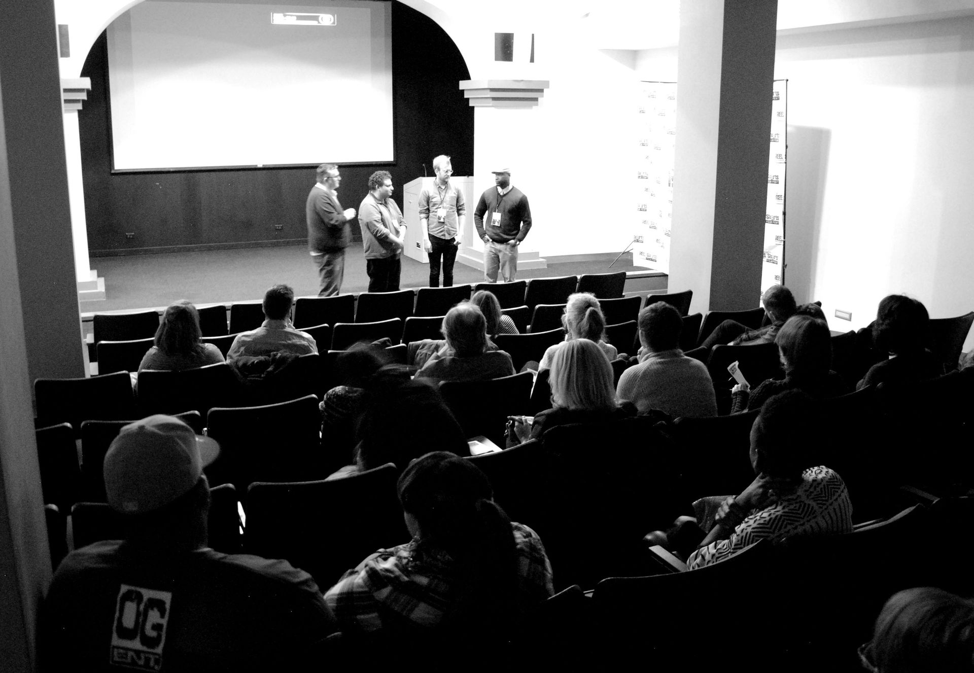 People in an auditorium listen to a presentation. Four speakers stand on a stage.