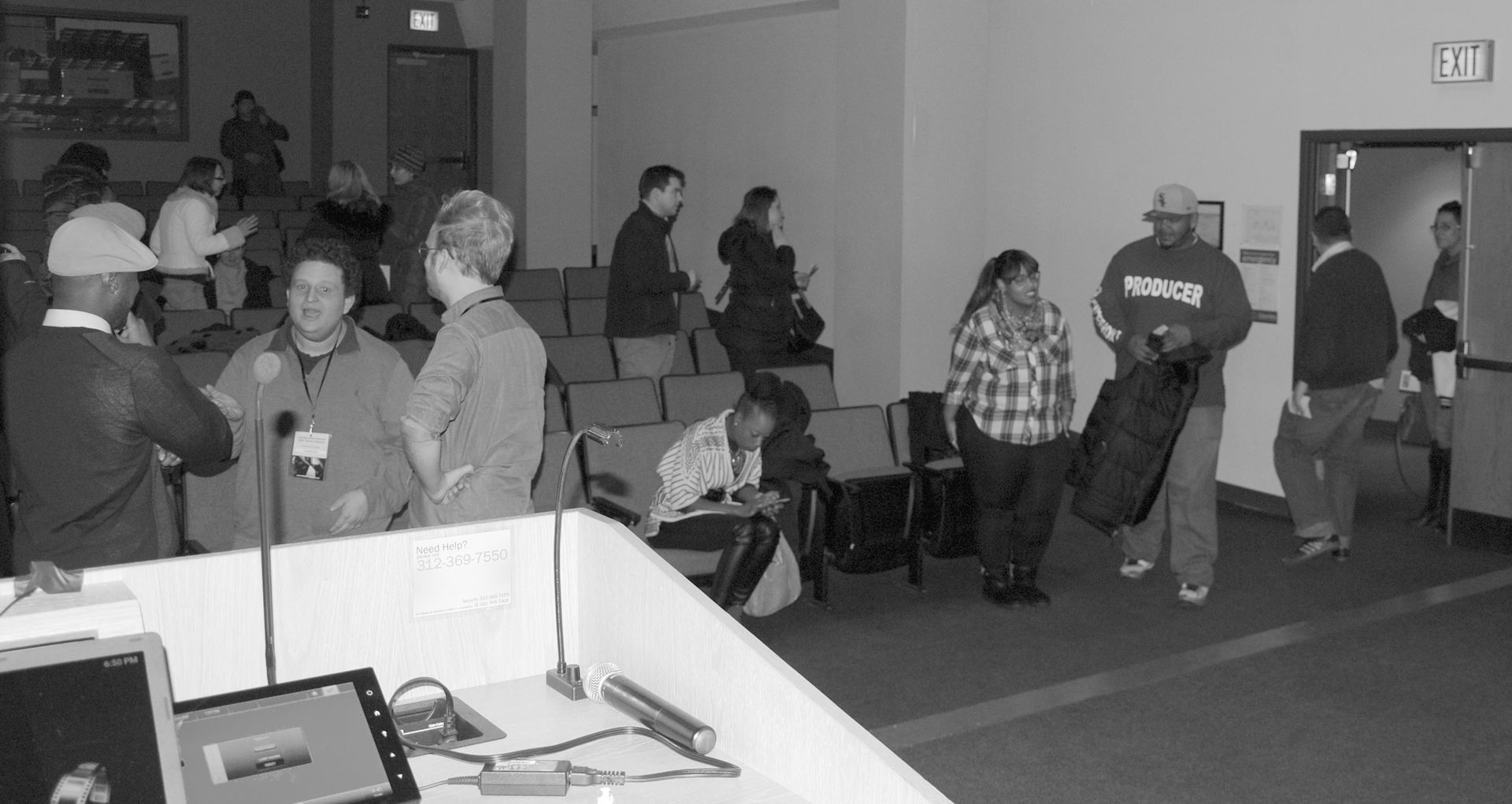 People gathered in a hall, some standing, others sitting. A man exits a door carrying a black bag.