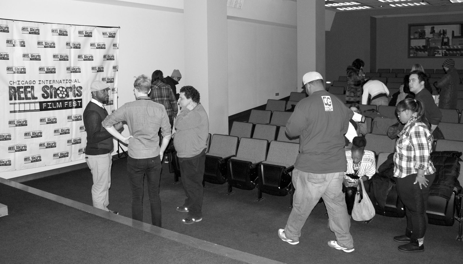 People gather in a theater lobby, some near a banner, others by seats. Black and white.