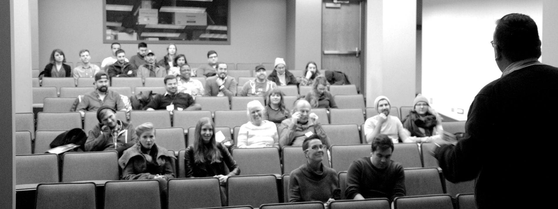 A lecture hall with rows of seated people, a presenter, and an artwork on the wall.