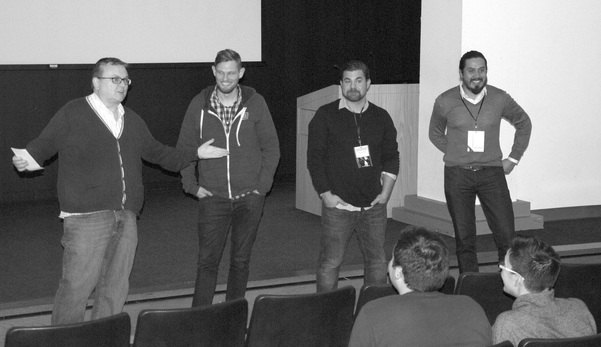 Four men stand on a stage, one gesturing and speaking. Audience members sit in chairs.