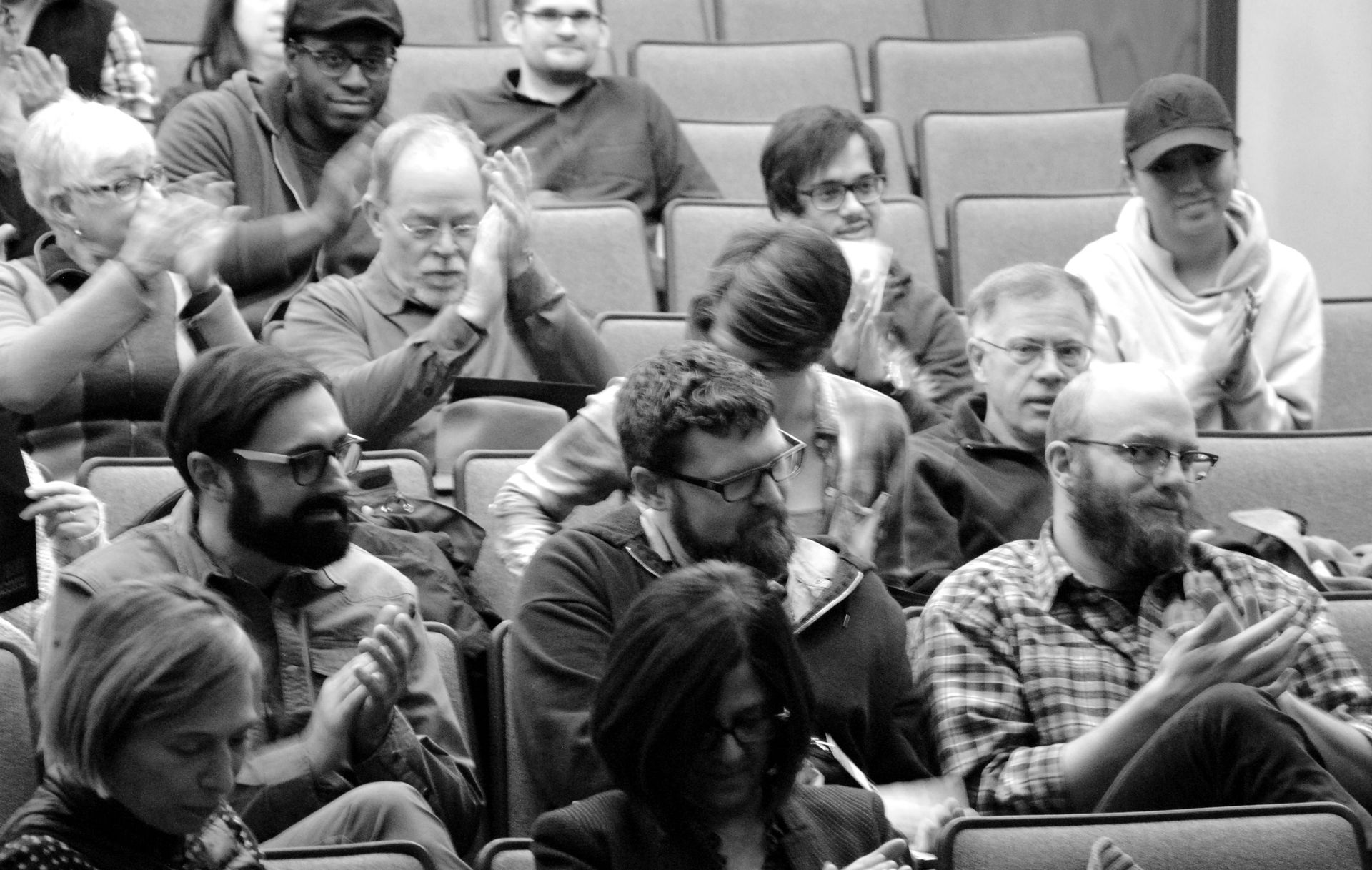 Audience clapping in an auditorium. People of various ages and genders watch, some with focused expressions. Black and white.