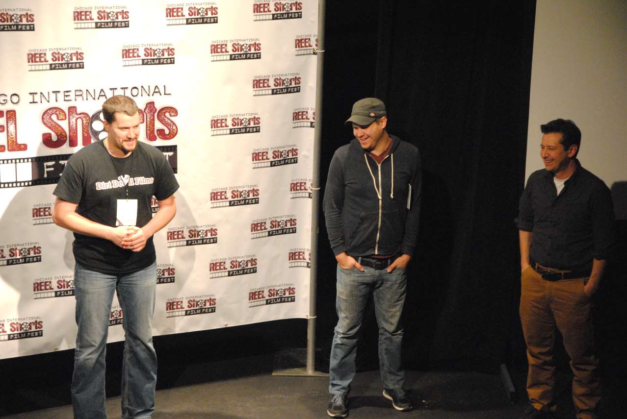 Three men stand on stage at a film festival, smiling. One wears a baseball cap and jeans.