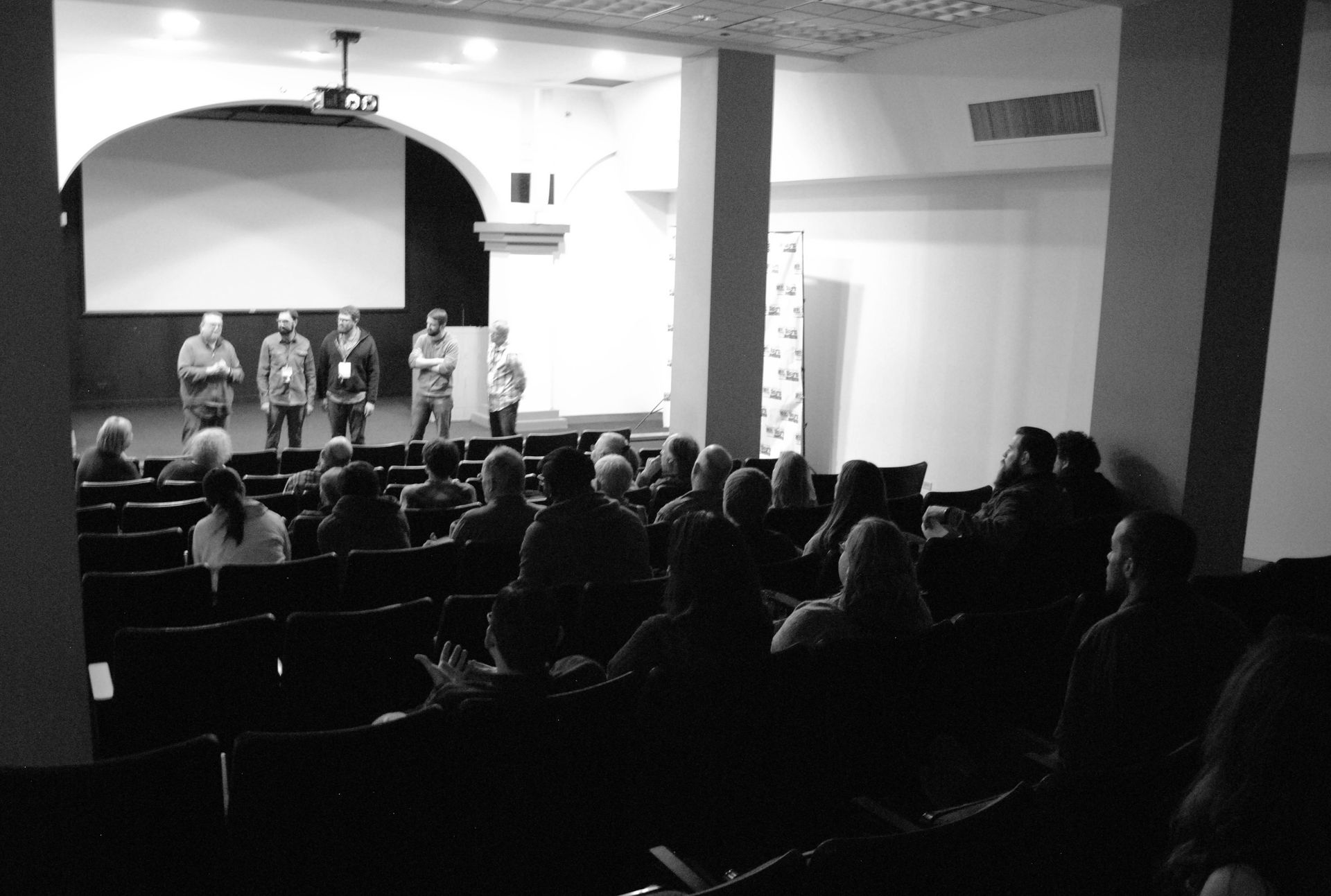 Audience in a theater watches speakers on stage, a screen above. Black and white.