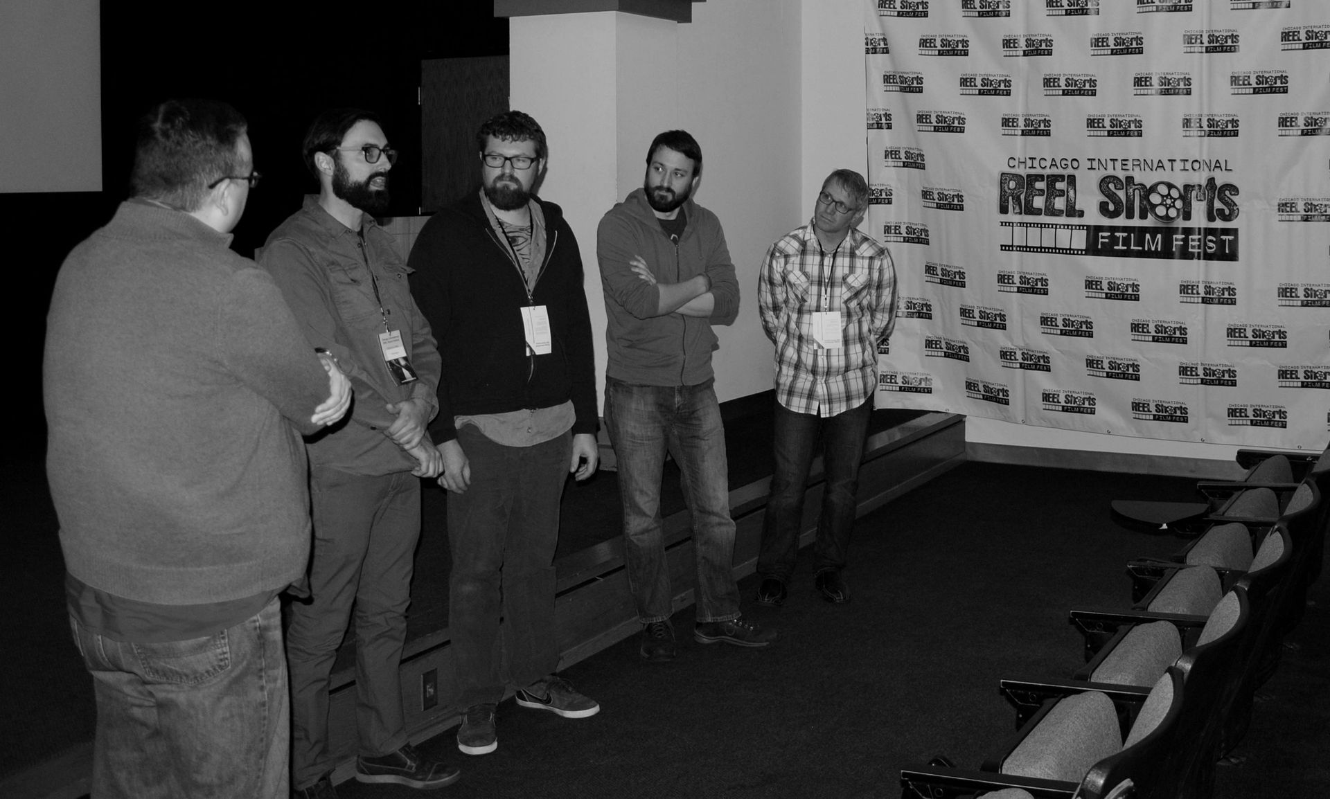 Five men stand in a room. One is facing the group with his arms crossed. The others are looking towards the right. A backdrop is visible.