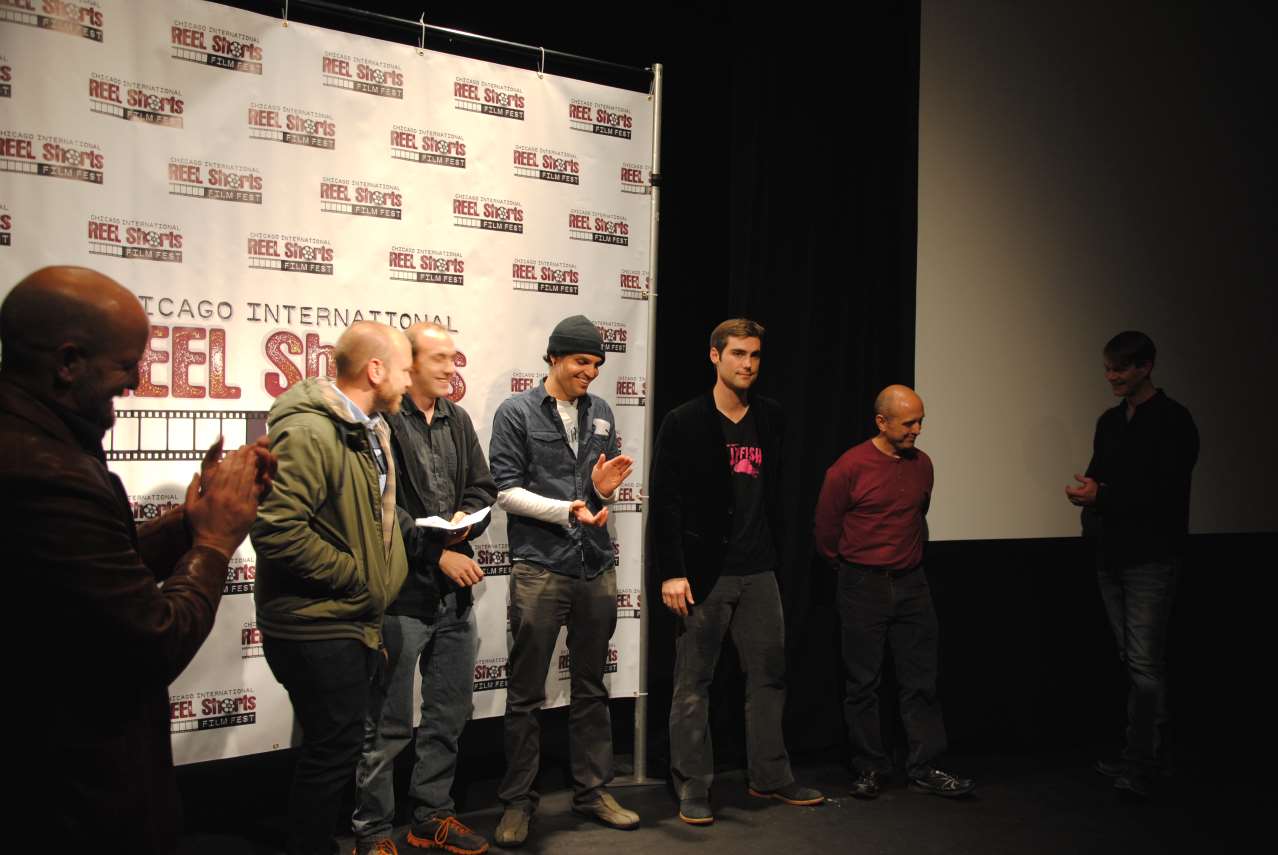 A group of people on a stage at an event. Men are holding an award and being applauded.