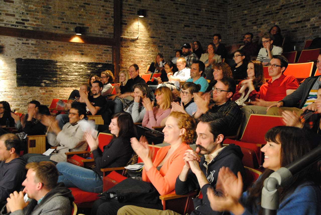 Audience applauds in a small theater. Red seats, brick wall, and warm lighting.