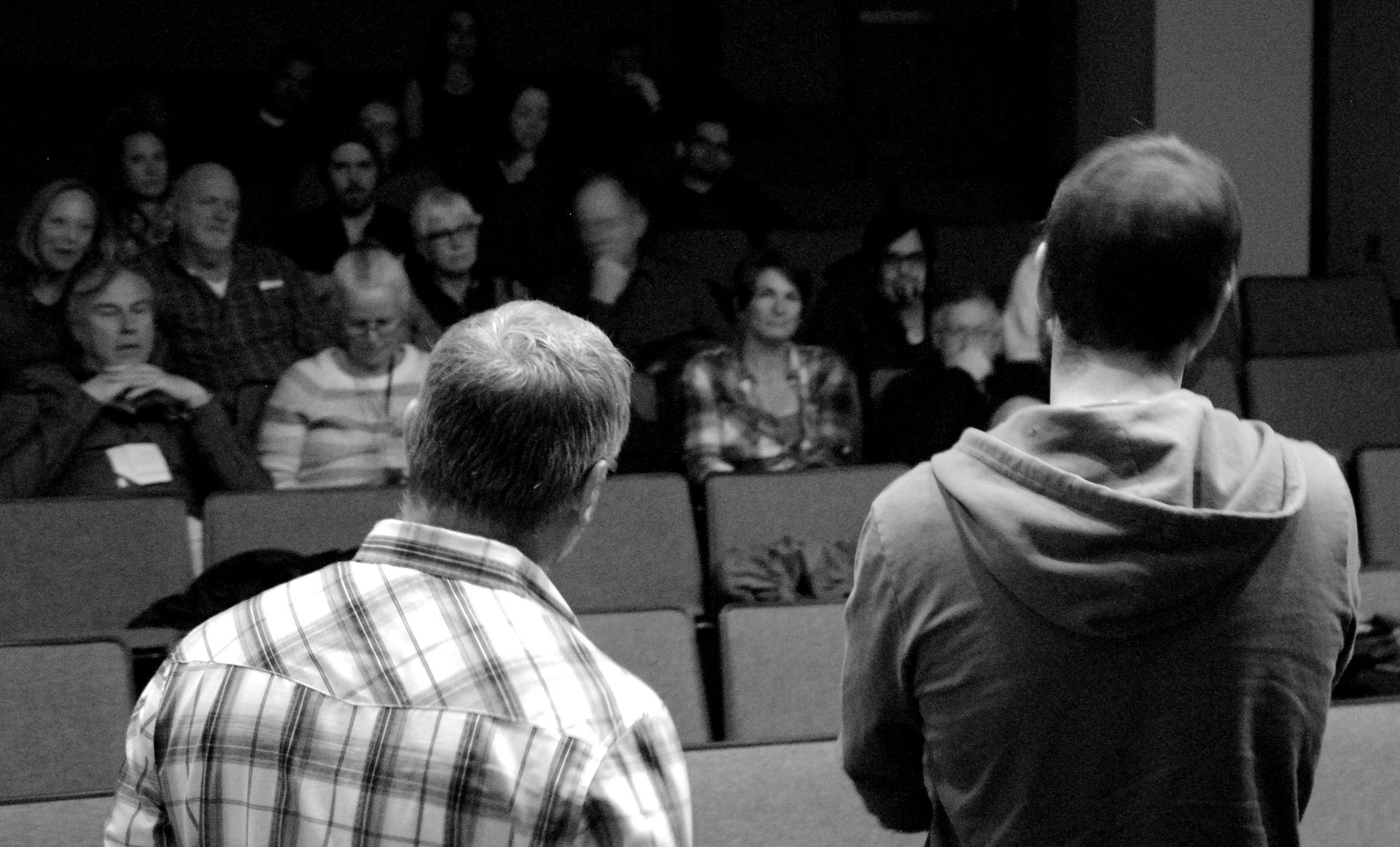 Two men face an audience in a theater. The audience is seated, watching.