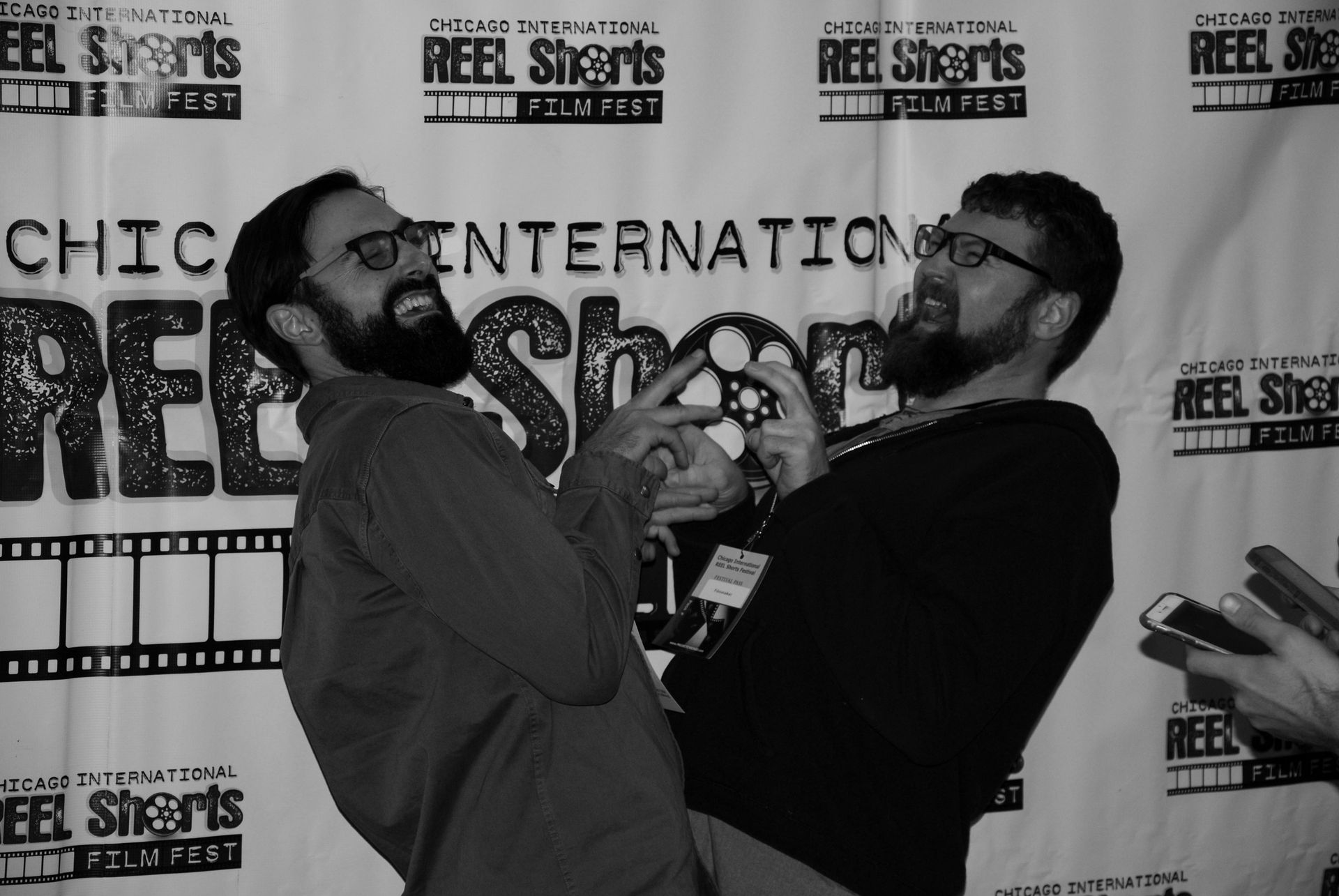 Two men with beards, glasses, and happy expressions, posing at a film festival, making hand gestures.