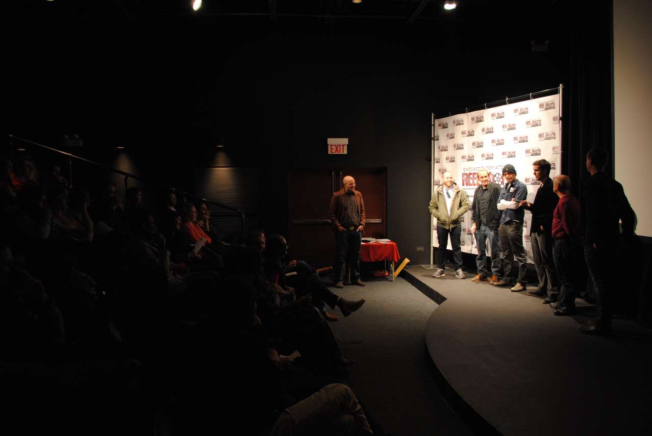 Audience in a dark movie theater watches a group on stage at a film premiere; a white backdrop is behind the group.