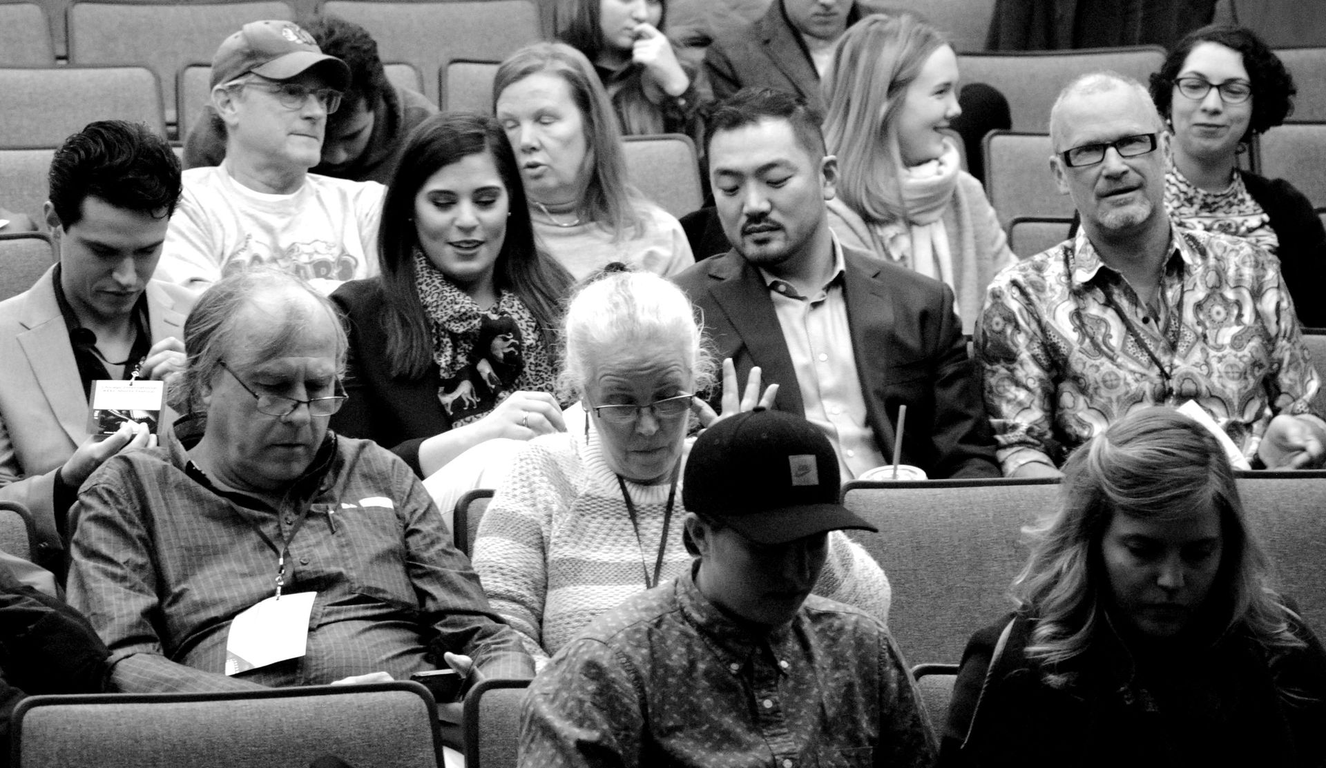 Audience members seated in an auditorium, some looking forward, others talking. Black and white photo.