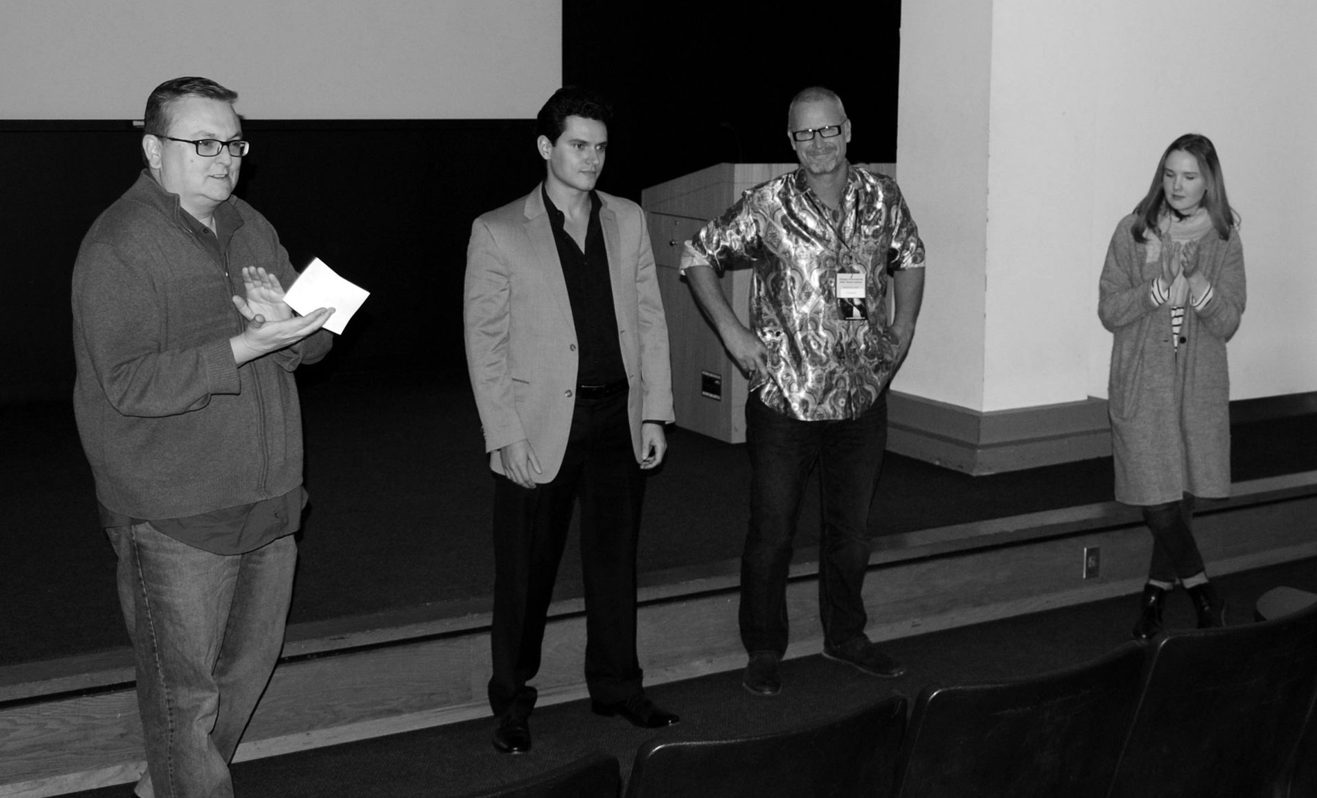 Four people on a stage: a man reading notes, two men and a woman standing. Black and white.