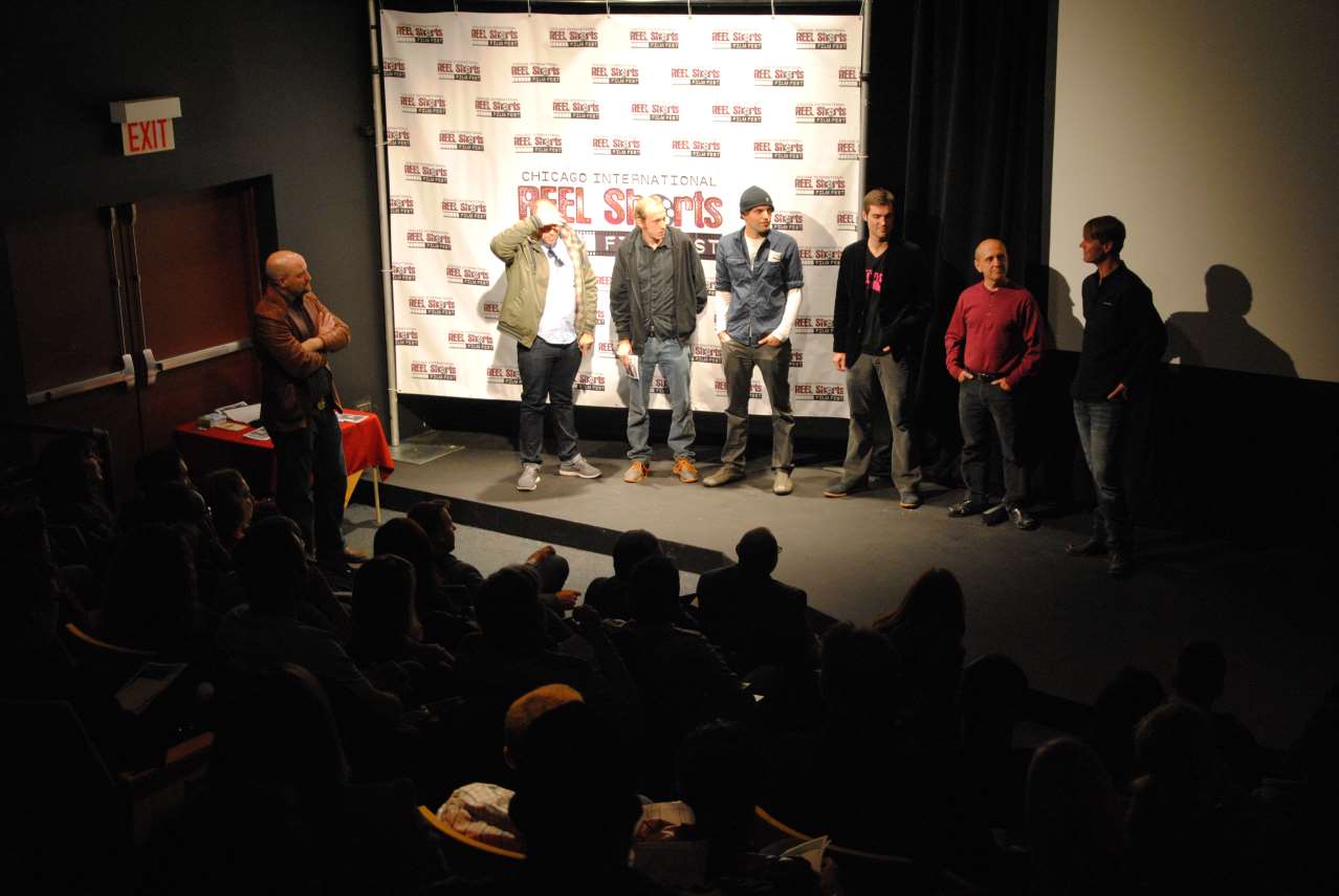 People on a stage in a theater, talking to audience. White backdrop behind them.