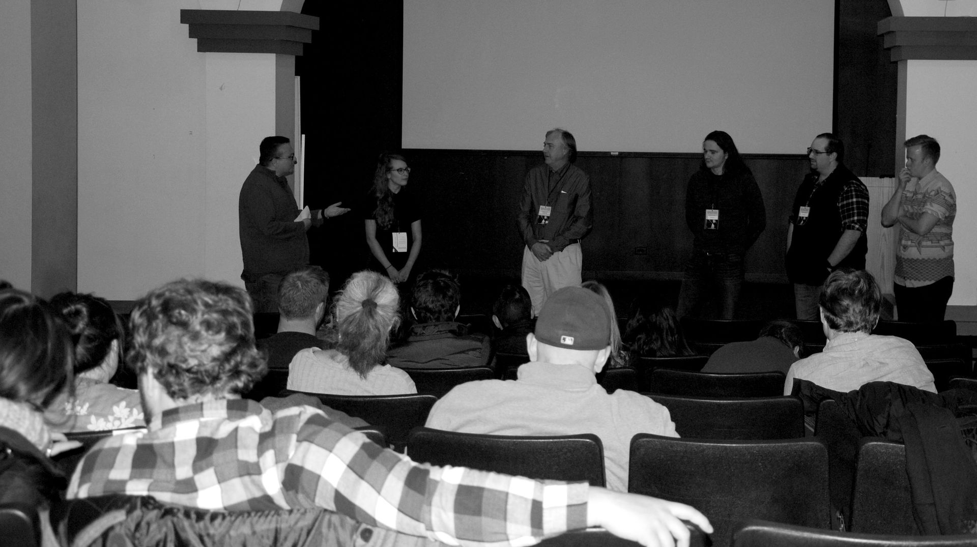Panel of speakers addressing audience in an auditorium. Speakers stand at the front. Audience faces them.