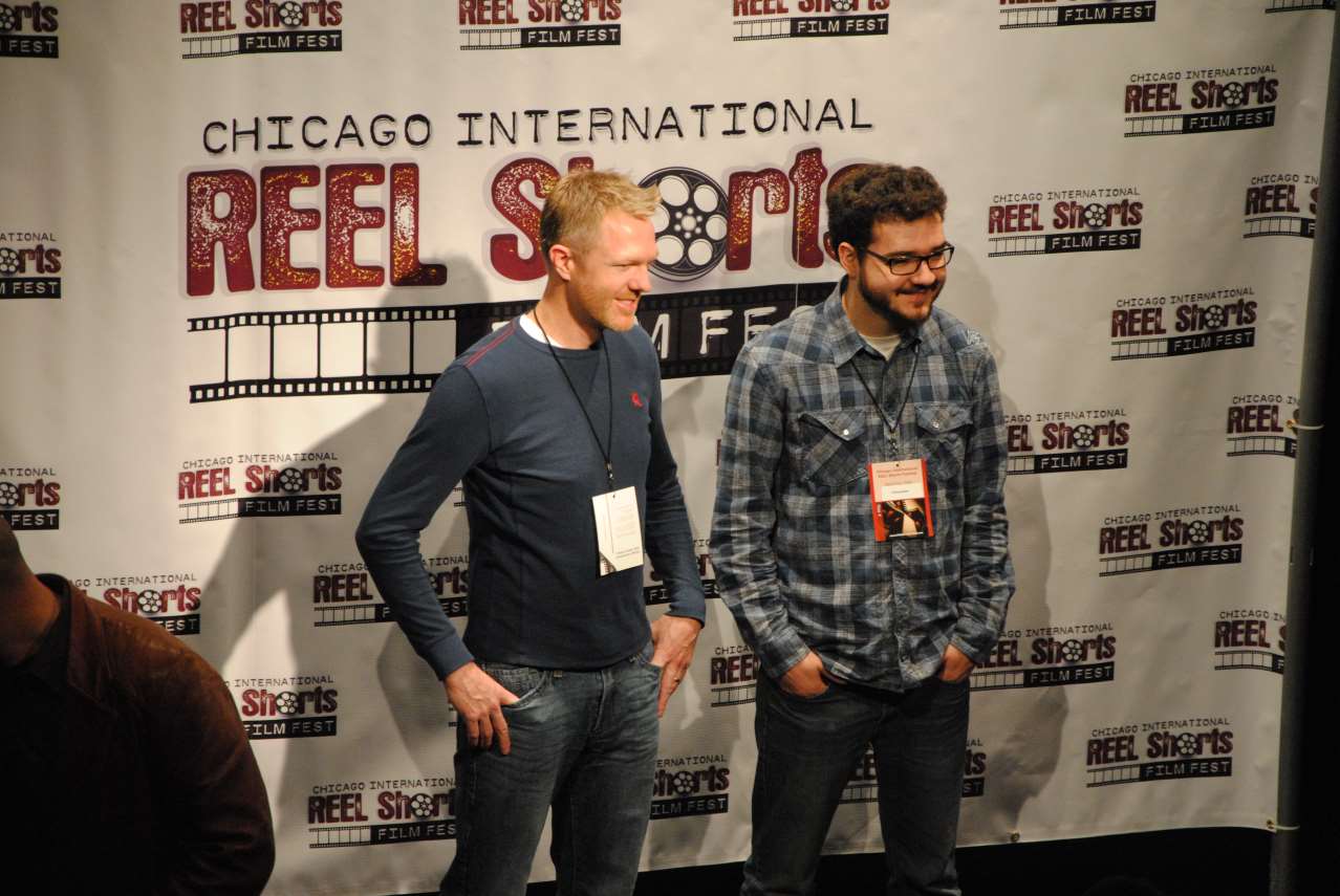 Two men smiling in front of a Chicago International Reel Shorts Film Fest backdrop; one in a blue shirt, one in a plaid shirt.