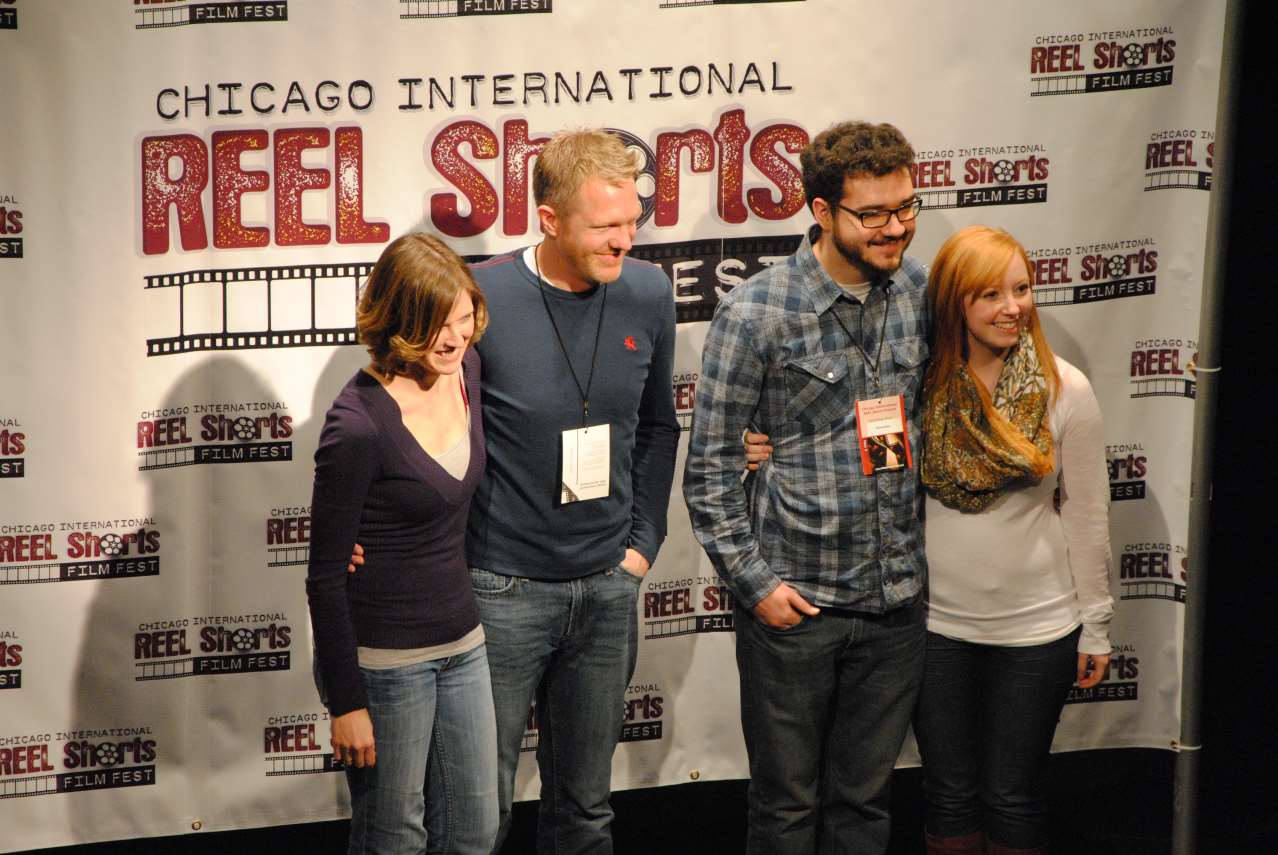 Four people smiling and posing at the Chicago International Reel Shorts Film Festival; a white backdrop.