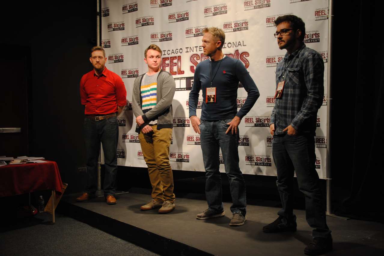 Four men stand on a stage in front of a banner. One in a red shirt, one in a striped sweater, one in a blue sweater, and one in a plaid shirt.