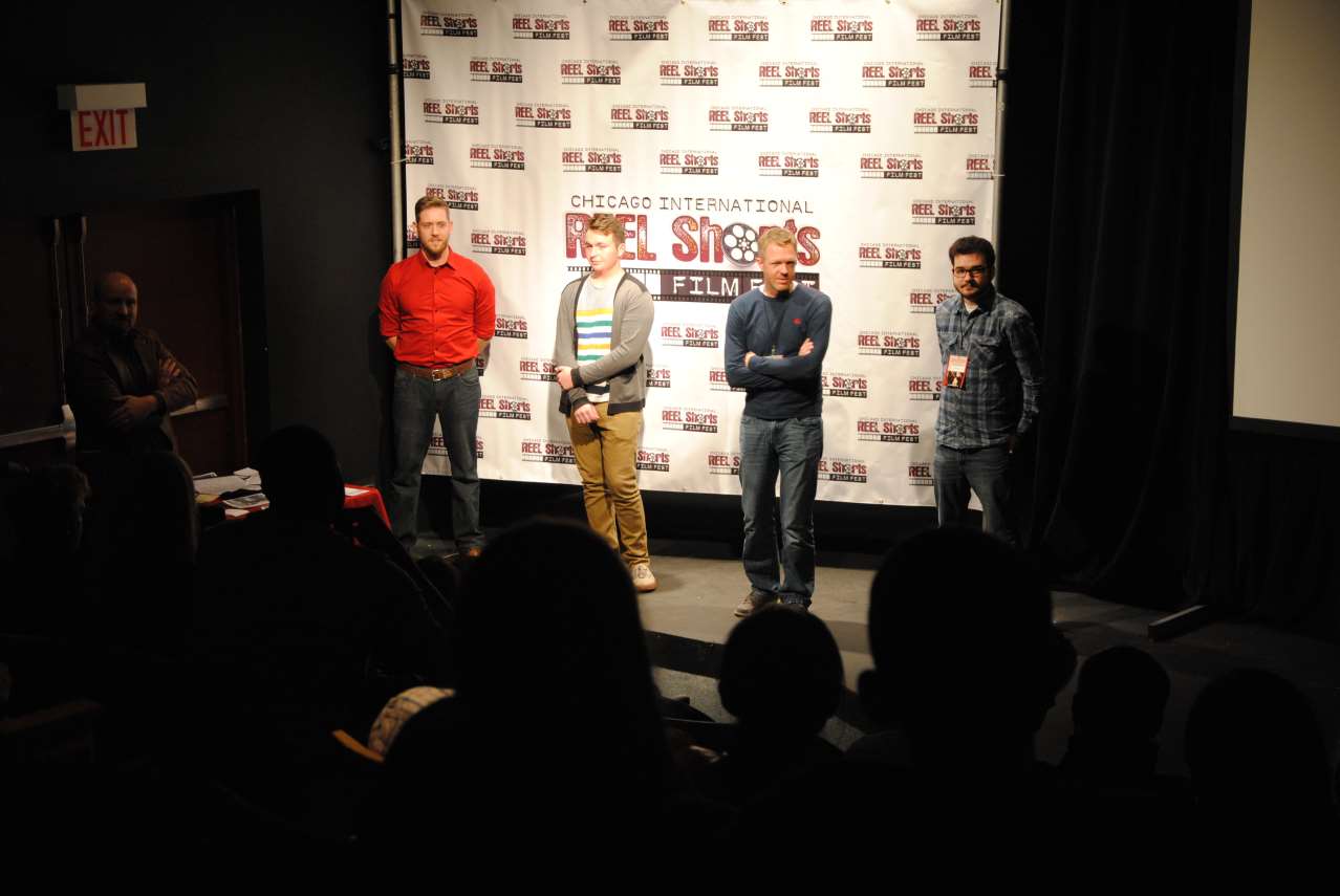 Men on stage at a film festival. Audience in foreground. Background has festival logo repeating.
