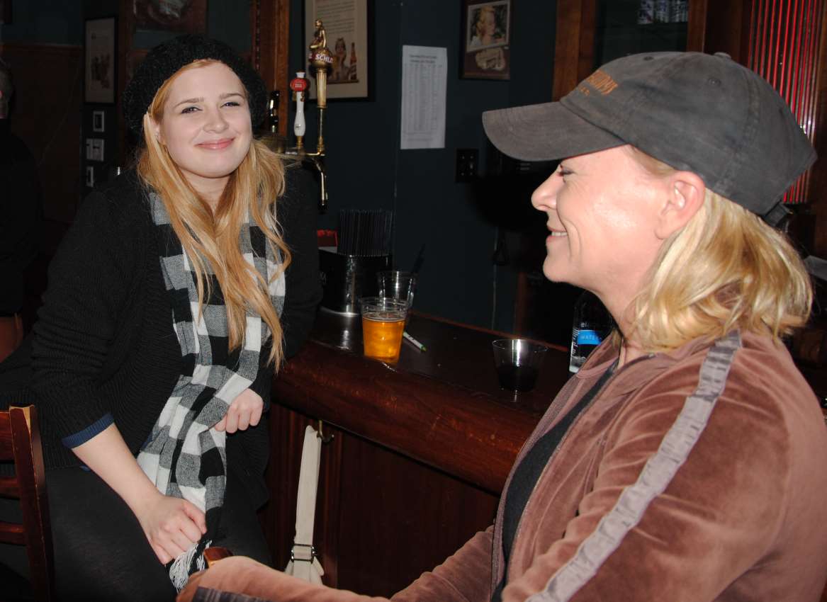 Two women at a bar. One smiles, wearing a beanie and scarf, the other smiles, wearing a baseball cap and brown jacket.