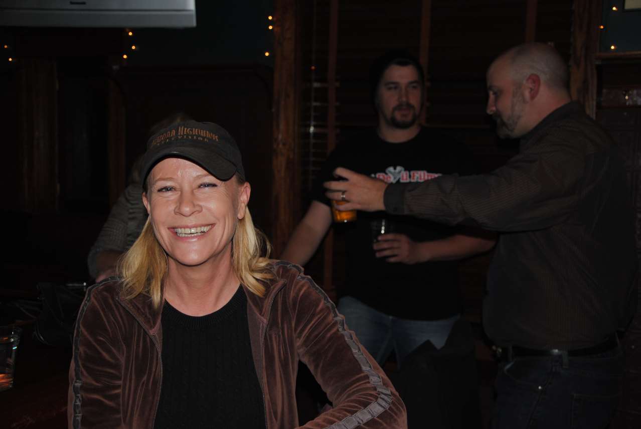 Woman in hat smiles at the camera, two men in background holding drinks, dark interior.
