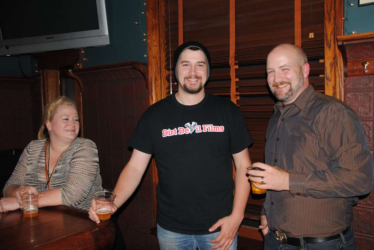 Three people smiling in a dimly lit bar. A woman at a table, a man in a black shirt, and a bald man holding a drink.