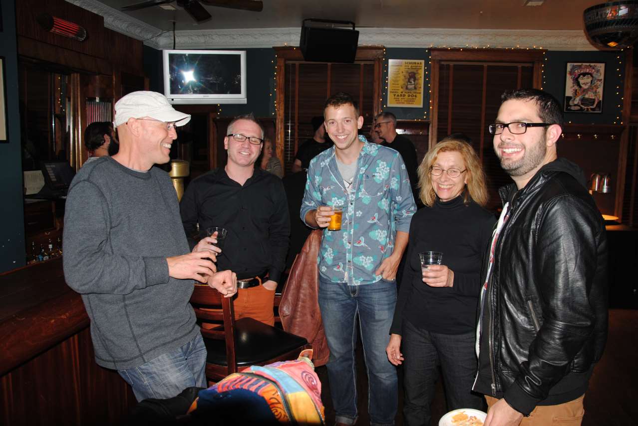 Five people smiling in a dimly lit bar; a man in a floral shirt holds a beer, others holding drinks.