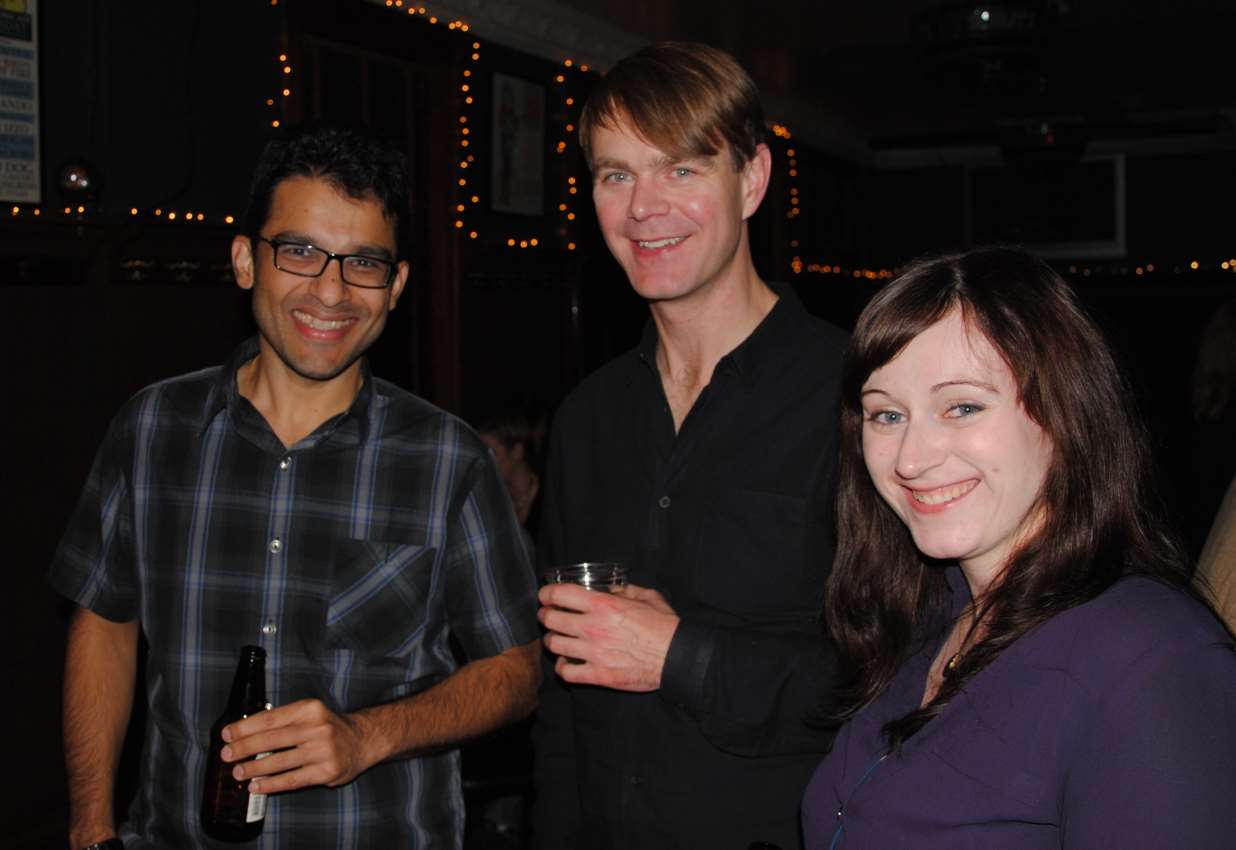 Three people smiling indoors: man in plaid, man in black, woman in purple. Dimly lit bar setting.
